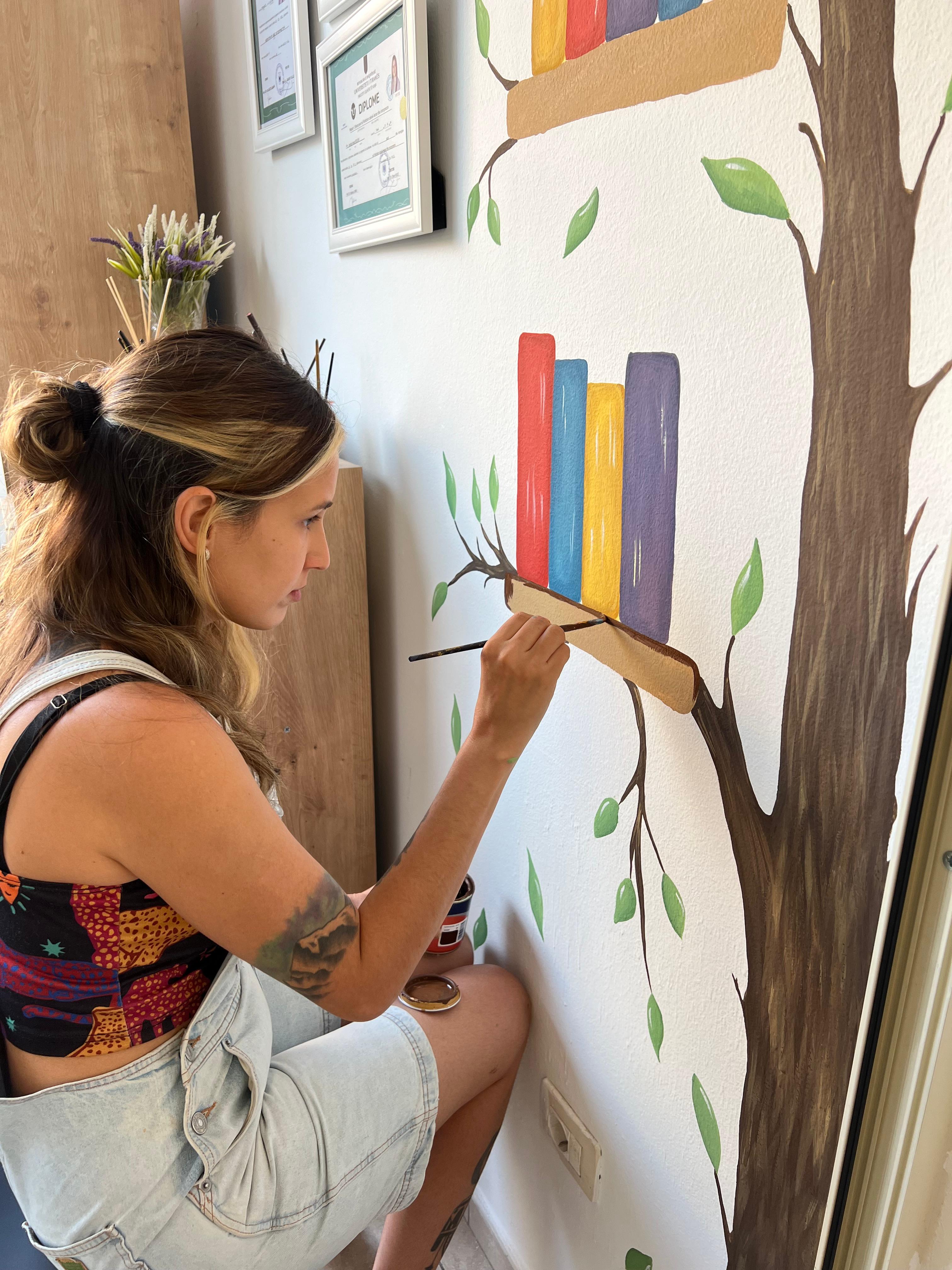 Paloma Kaiper hand-painting a mural of a tree and books