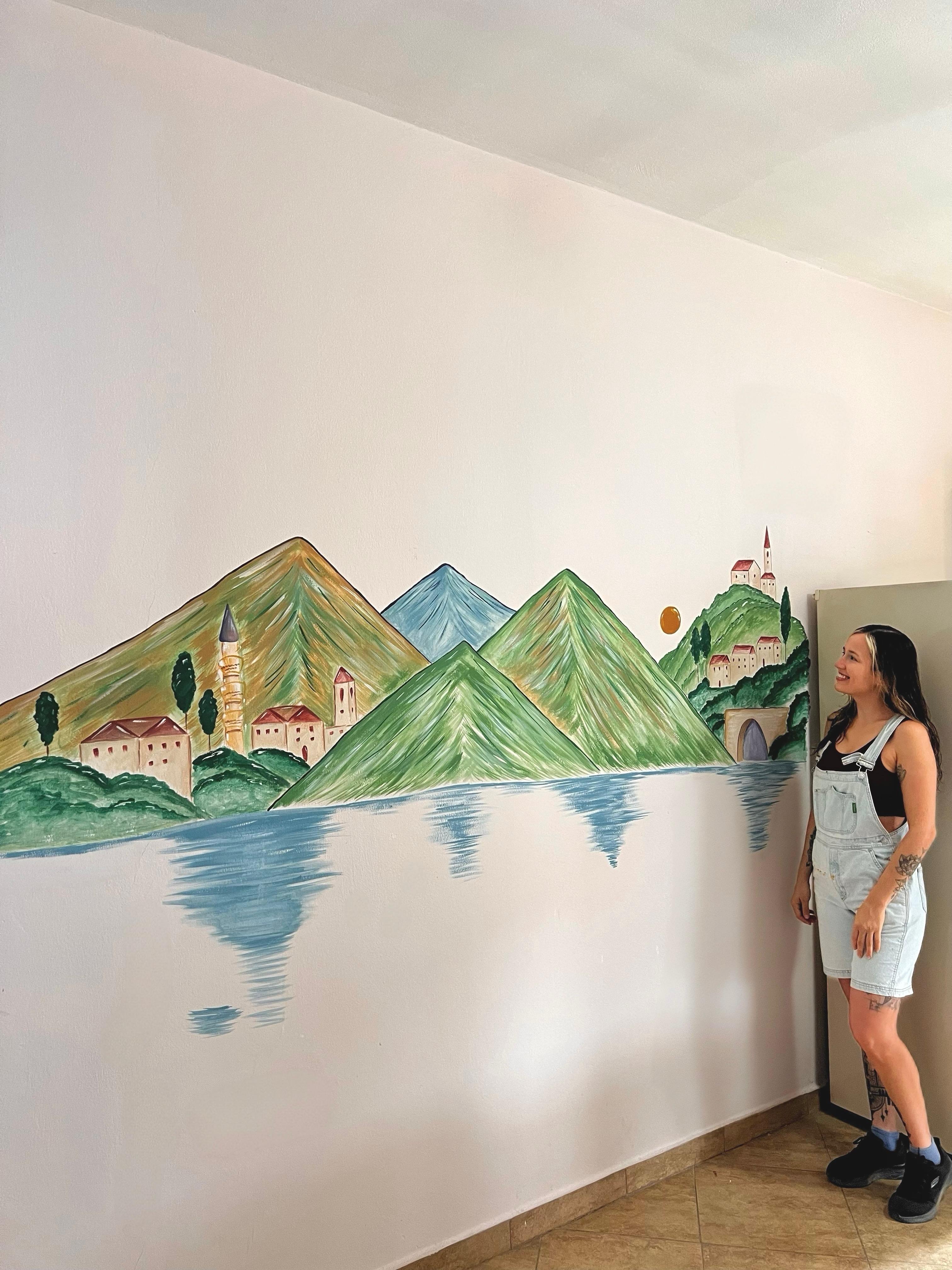 Paloma standing beside her mural of mountains, villages and a lake
